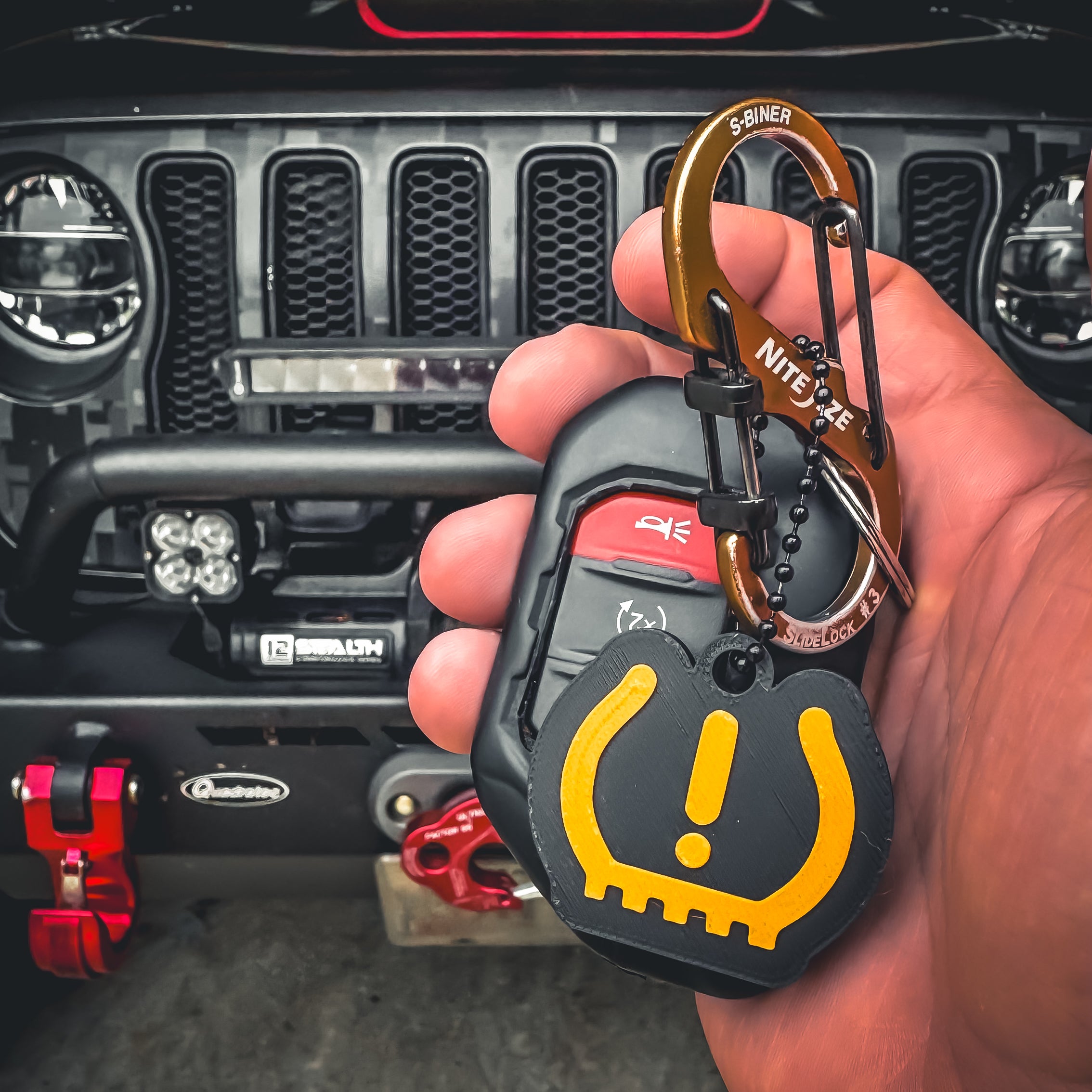 A hand holds Jeep car keys with the FirstWarrior Tire Pressure Keychain, a 3D printed yellow Jeep dash accessory. The background shows the front grille and bumper of a Jeep vehicle.