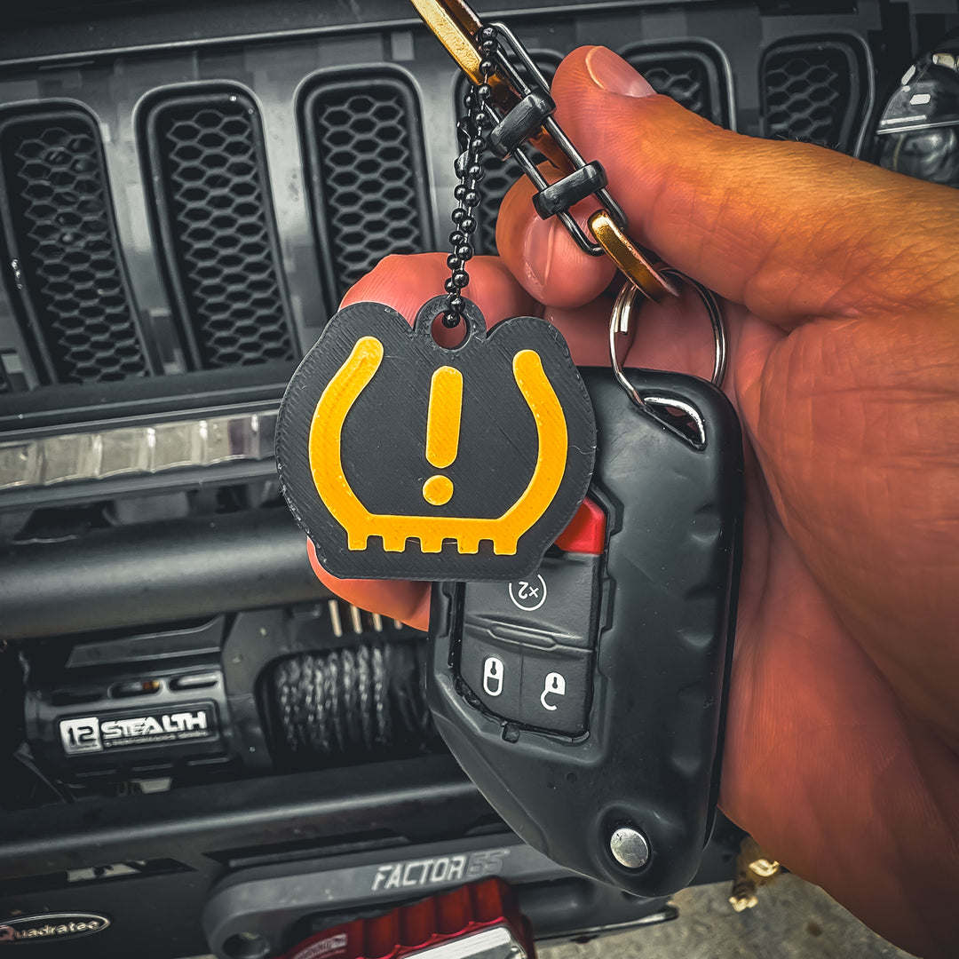 A hand holds a car key fob with the FirstWarrior Tire Pressure Keychain—3D printed in yellow PLA as a tire pressure warning symbol—an ideal Jeep dash accessory, shown before a vehicle grill and winch.