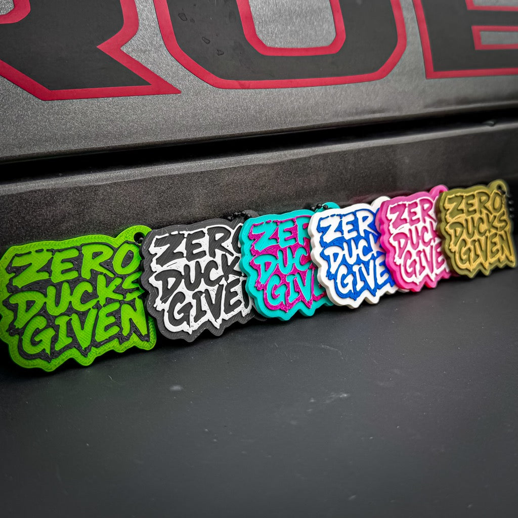Six vibrant "Zero Ducks Given" keychains by FirstWarrior are displayed on a black and red surface—great for your car dashboard or paired with a 3D-printed duck. Each keychain features bold color combos like green, black, and pink.