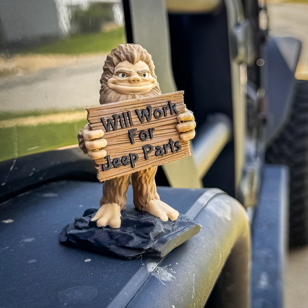 The FirstWarrior Will Work for Parts! Bigfoot Deskmate is a small PLA figurine for your Jeep, holding a wooden "Will Work for Parts" sign on a black Jeep bumper, with part of the vehicle and tire visible in the background.