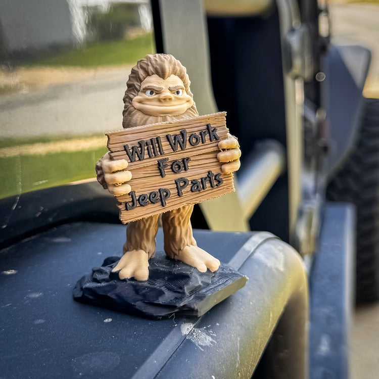 The FirstWarrior Will Work for Parts! Bigfoot Deskmate is a small PLA figurine for your Jeep, holding a wooden "Will Work for Parts" sign on a black Jeep bumper, with part of the vehicle and tire visible in the background.