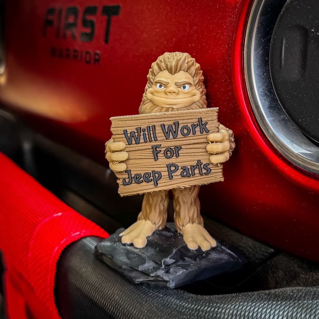 The FirstWarrior Will Work for Parts! Bigfoot Deskmate is a small PLA jeep accessory figurine holding a "Will Work For Jeep Parts" sign, designed to sit near the dashboard and air vent of your red Jeep.