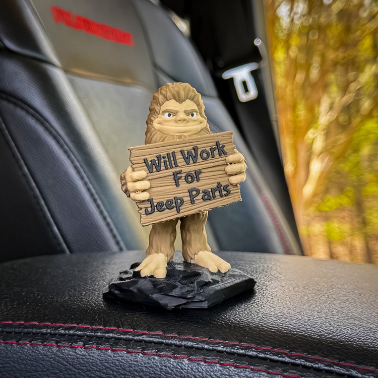 The FirstWarrior Will Work for Parts! Bigfoot Deskmate, a quirky PLA plastic figurine, sits on a black car seat with red stitching, holding its signature sign and framed by trees seen through the window.