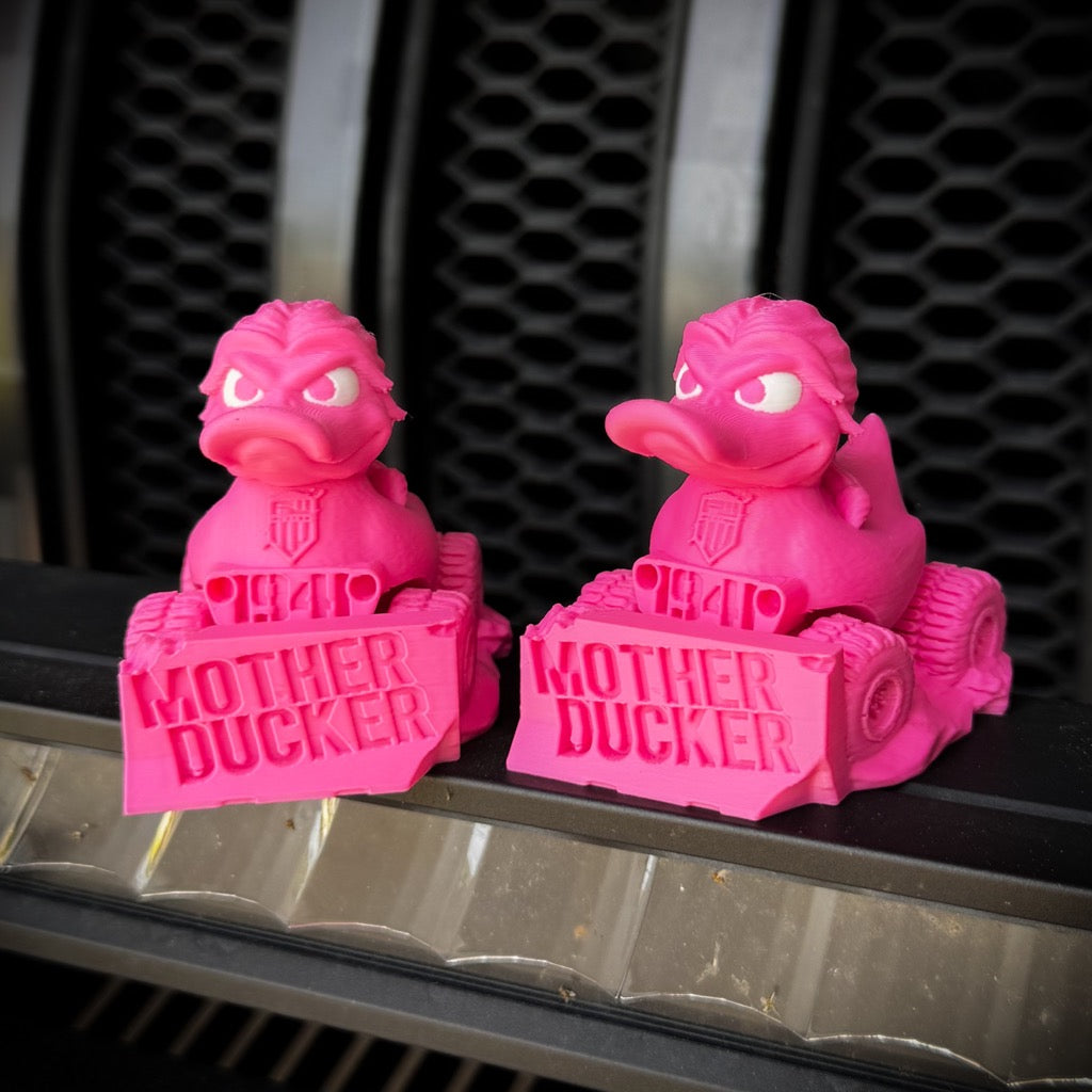 Two bright pink rubber ducks with fierce expressions sit in toy bulldozers labeled MOTHER DUCKER and 1941. These limited edition Mother Ducker - v2.0 figures by FirstWarrior are made of PLA plastic and set against a metallic grill-like background.