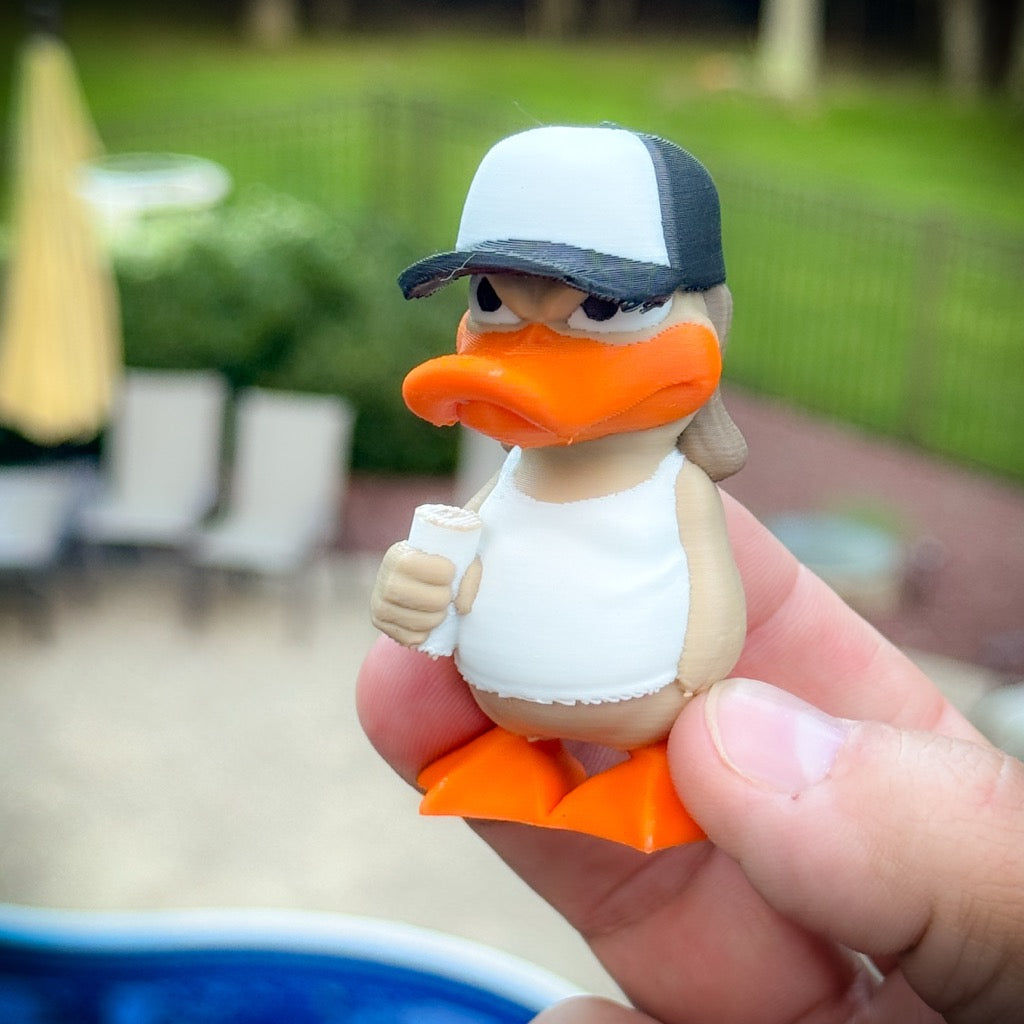 A hand holds Bubba Duck, a small 3D-printed figure by FirstWarrior, in a white tank top and black-and-white cap, clutching a rolled item, with a blurred backyard and pool in the background.