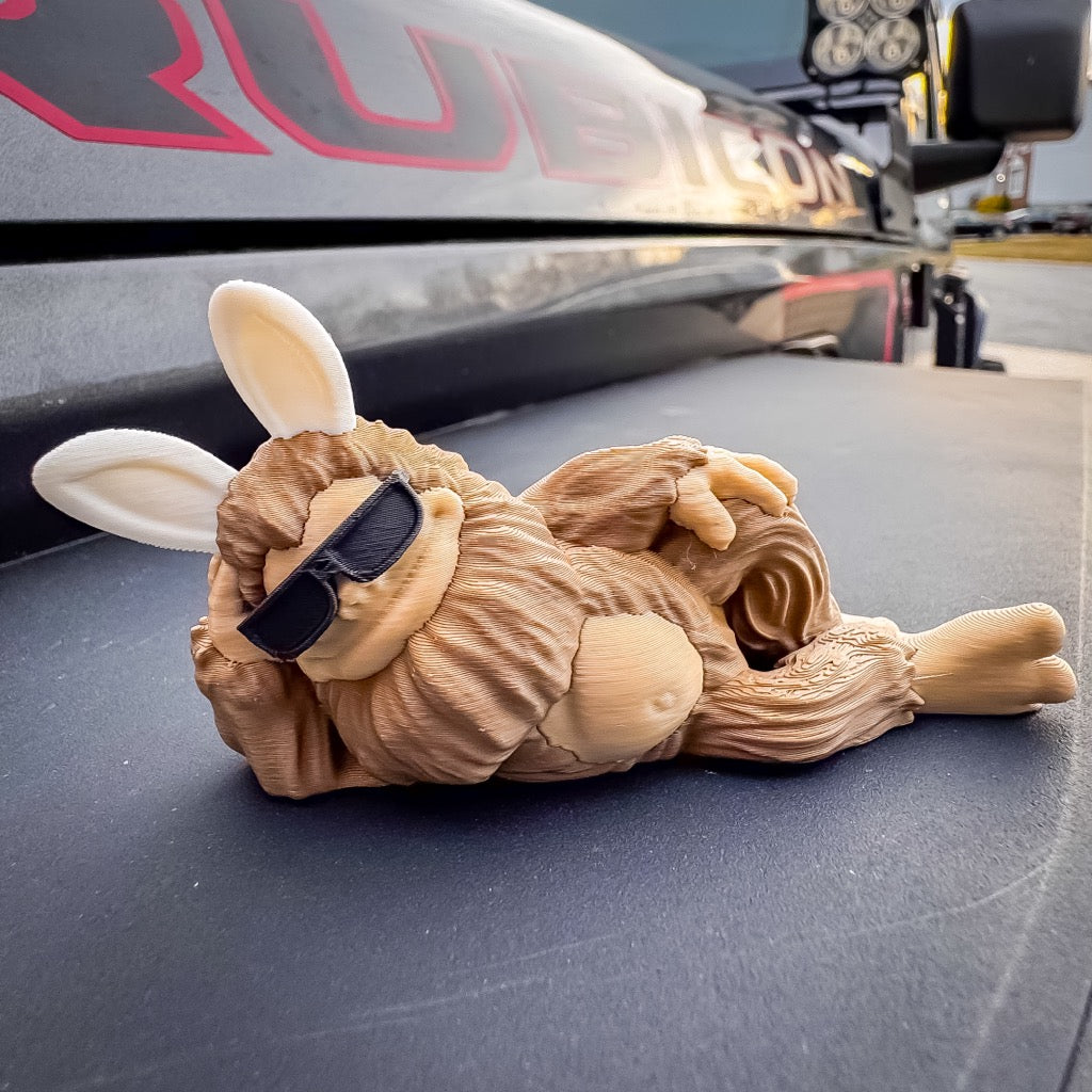 A small, furry Easter Imposter Bigfoot Limited Edition Deskmate by FirstWarrior, featuring bunny ears and sunglasses, lounges on its side—making it a quirky car dashboard decoration.