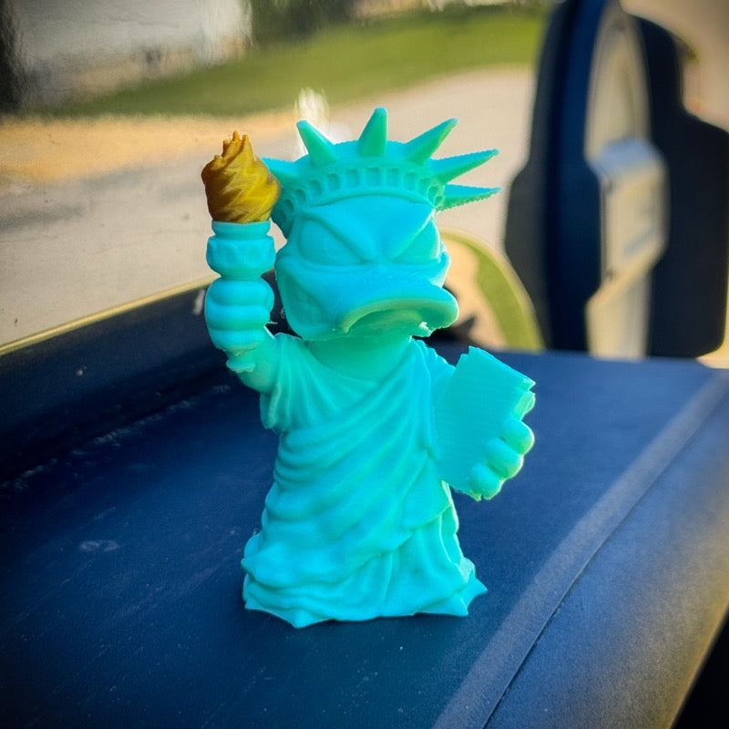Turquoise Statue of Liberty figurine on a car dashboard with a blurred outdoor background