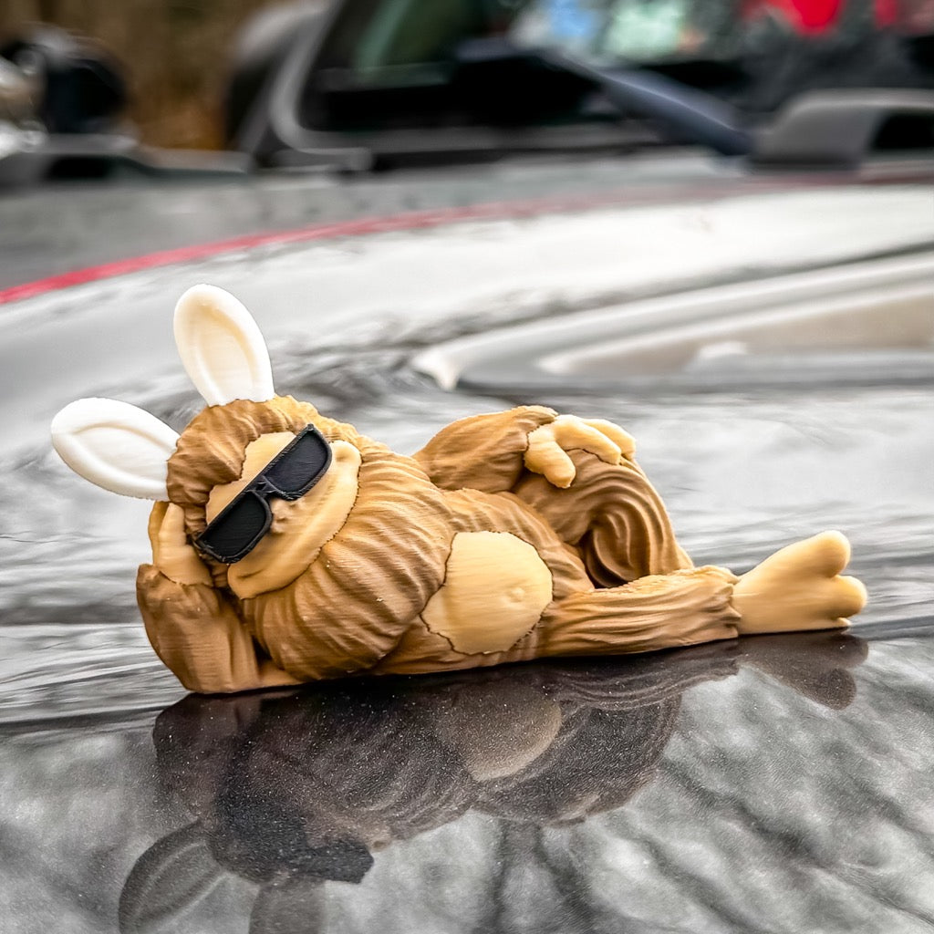 The FirstWarrior Easter Imposter Bigfoot Limited Edition Deskmate, featuring bunny ears and sunglasses, lounges atop a car hood as a quirky dashboard decoration, with blurred cars in the background.