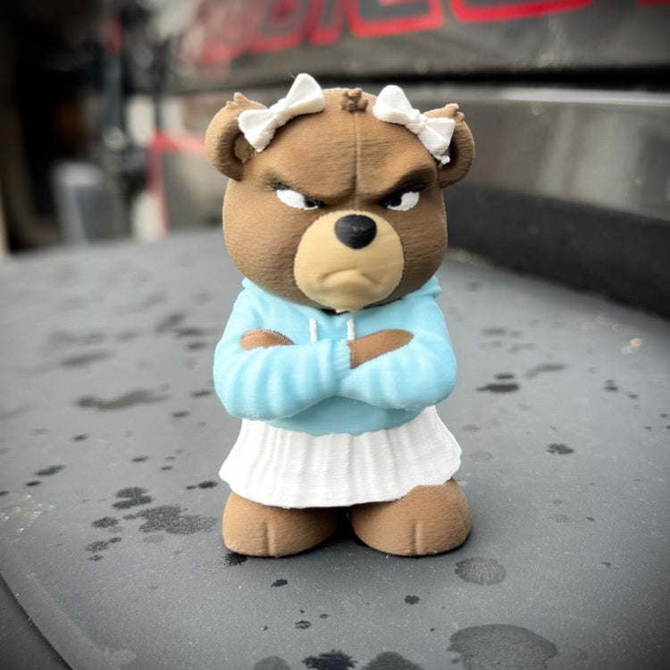 Stuffed bear toy with an angry expression wearing a blue sweater and white skirt on a car hood.