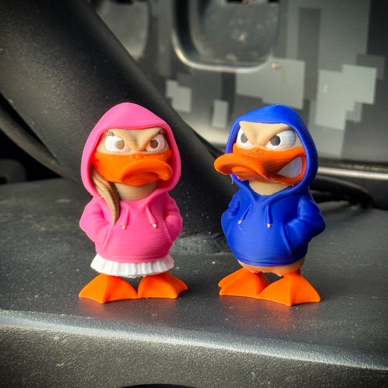 Two duck figurines wearing pink and blue hoodies on a car dashboard.