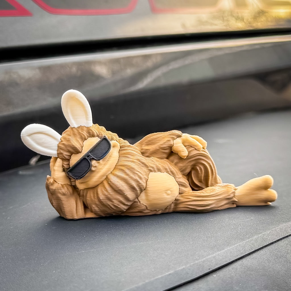 The FirstWarrior Easter Imposter Bigfoot Limited Edition Deskmate, featuring bunny ears and sunglasses, lounges playfully—an adorable choice for quirky car dashboard decor.