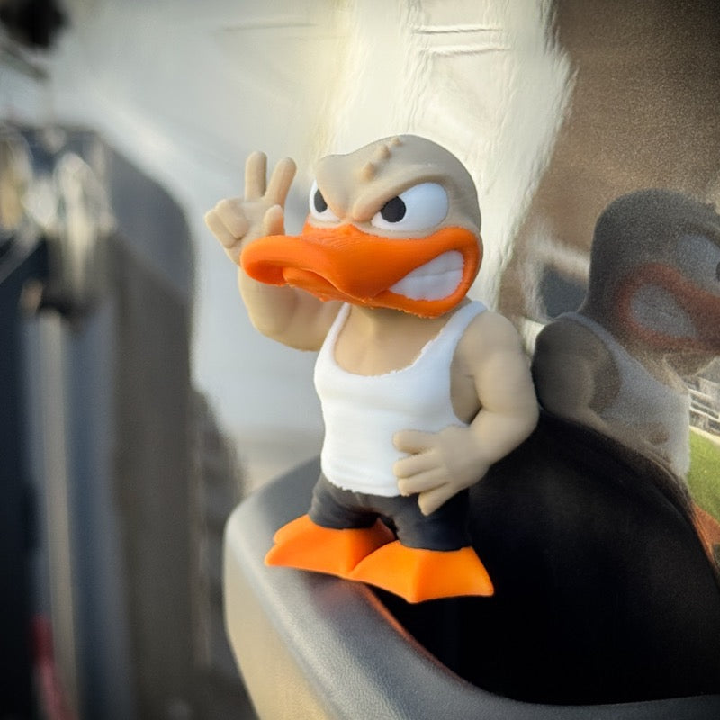 The FirstWarrior Tough Guy Duck figurine, crafted from PLA plastic, sports an angry look with a white tank top and black pants, flashing a peace sign—an ideal quirky accessory for your car dashboard with its reflection shining behind it.