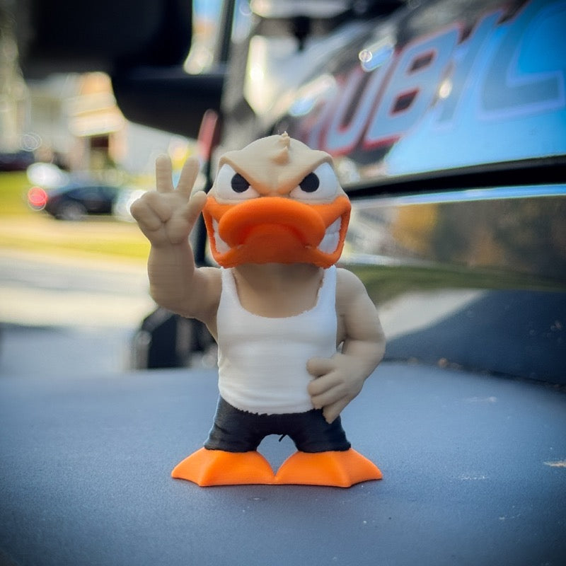 The FirstWarrior Tough Guy Duck, a small PLA dashboard accessory, stands outdoors in a white tank top and black shorts, one hand on its hip and the other showing a peace sign by a shiny, dark surface.
