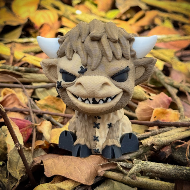The FirstWarrior Patches Baby Highland—a small desk ornament with horns, dark eyes, sharp teeth, and stitched details—sits among autumn leaves and sticks on the ground.