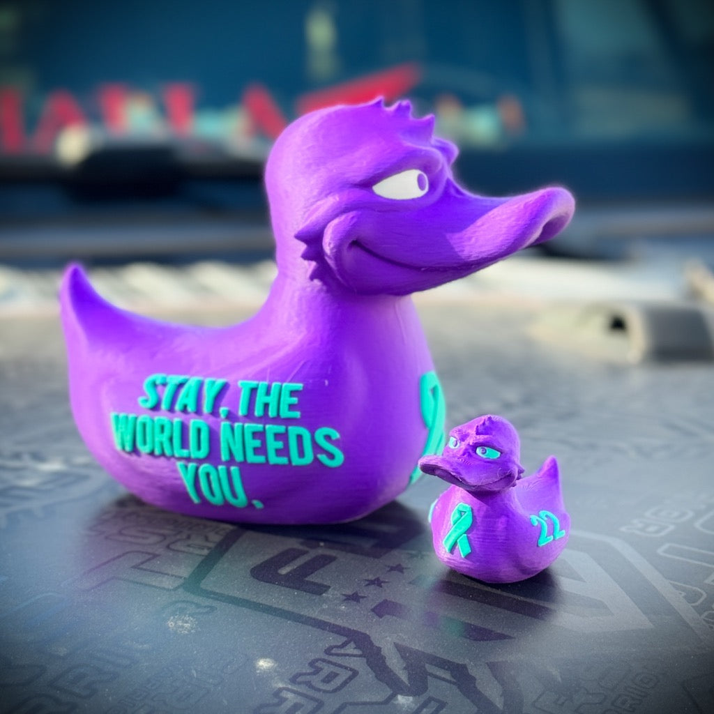 Two purple PLA ducks from FirstWarrior’s "22 a Day - The World Needs You" sit on a car hood. Both feature teal writing, teal ribbons, and the number 22 to promote veteran suicide prevention awareness.
