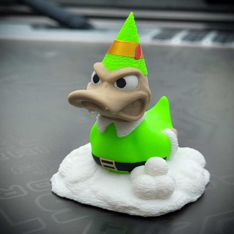 Buddy the Duck by FirstWarrior is a small figurine in a green Christmas elf outfit and party hat, standing on a white cloud-like base with two snowballs—perfect as festive decor or a fun car dashboard accessory.