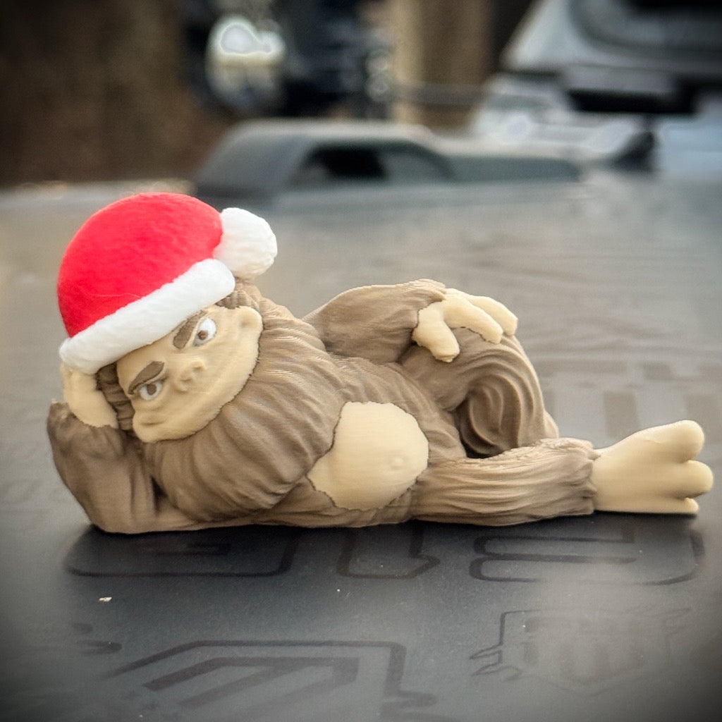 The FirstWarrior Hunky Claus Bigfoot figurine lounges in a red and white Santa hat, making it a fun Christmas gift or unique Jeep accessory.