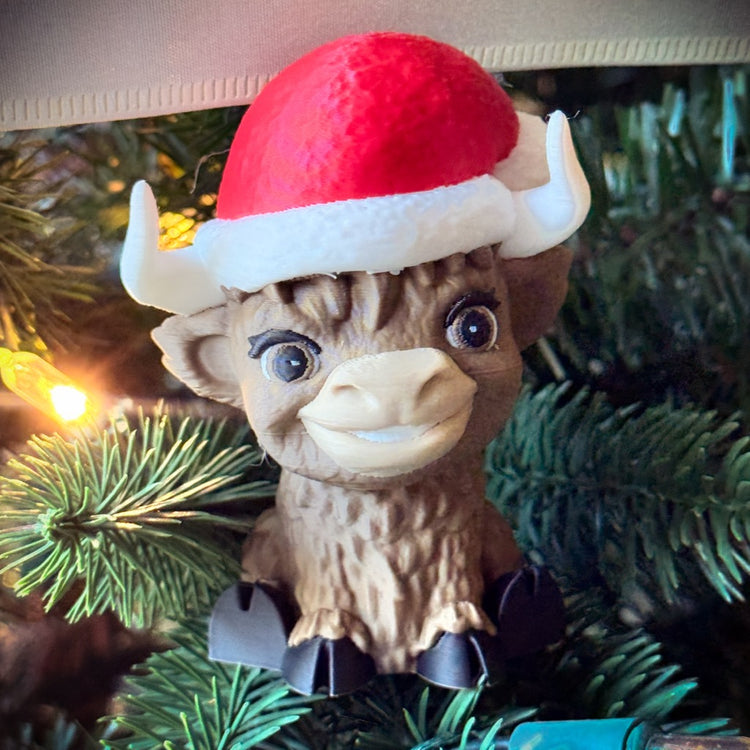 The FirstWarrior Baby Claus Baby Highland ornament, a cute brown buffalo resembling a baby Highland cow in a red and white Santa hat, sits nestled among Christmas tree pine needles and holiday lights.
