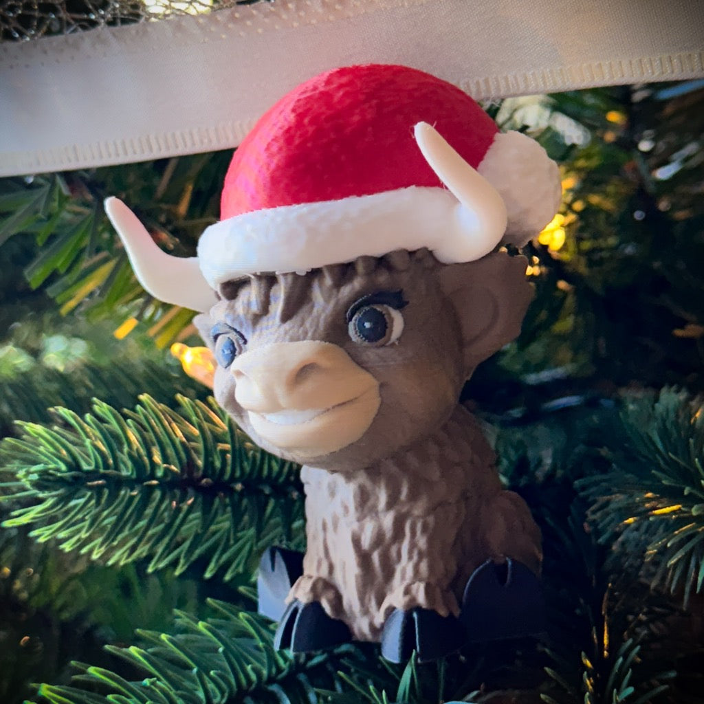 The FirstWarrior Baby Claus Baby Highland is a small smiling Highland cow figurine in a red Santa hat, nestled among pine branches with glowing yellow lights—ideal as a Jeep dashboard accessory or unique 3D-printed desk decor.