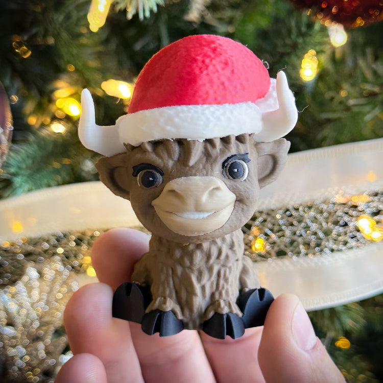 Someone holds a FirstWarrior "Baby Claus Baby Highland"—a cute, brown Highland buffalo figurine with a red and white Santa hat, ideal as Jeep dashboard decor or 3D-printed desk accessory—in front of a lit, ribbon-trimmed Christmas tree.