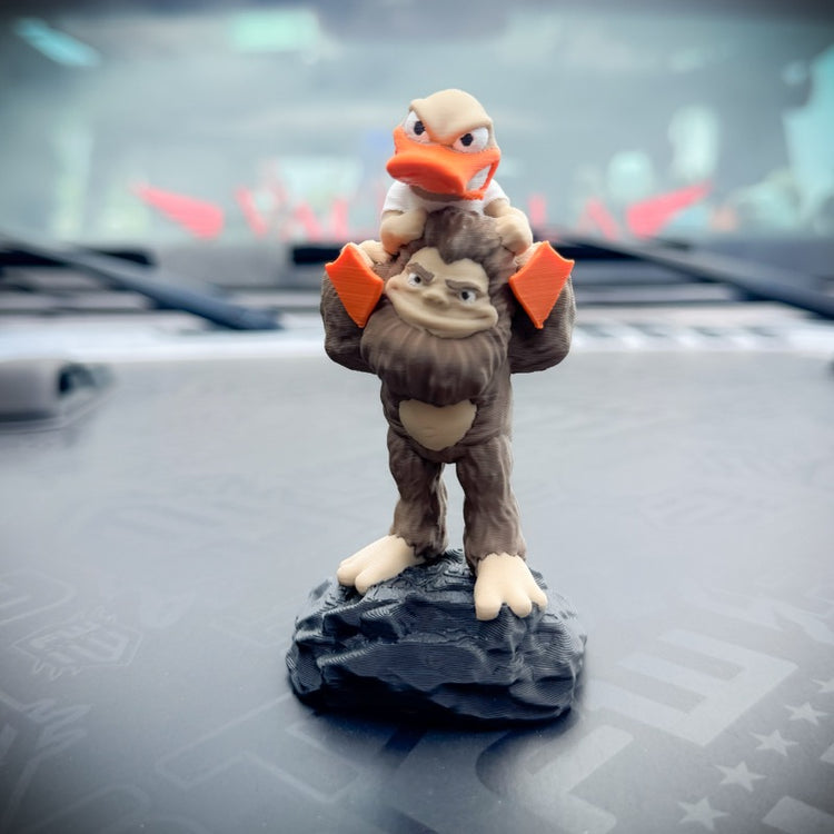 A small figurine of a brown bigfoot carrying a duck on its shoulders, mounted on a dark gray rock base,