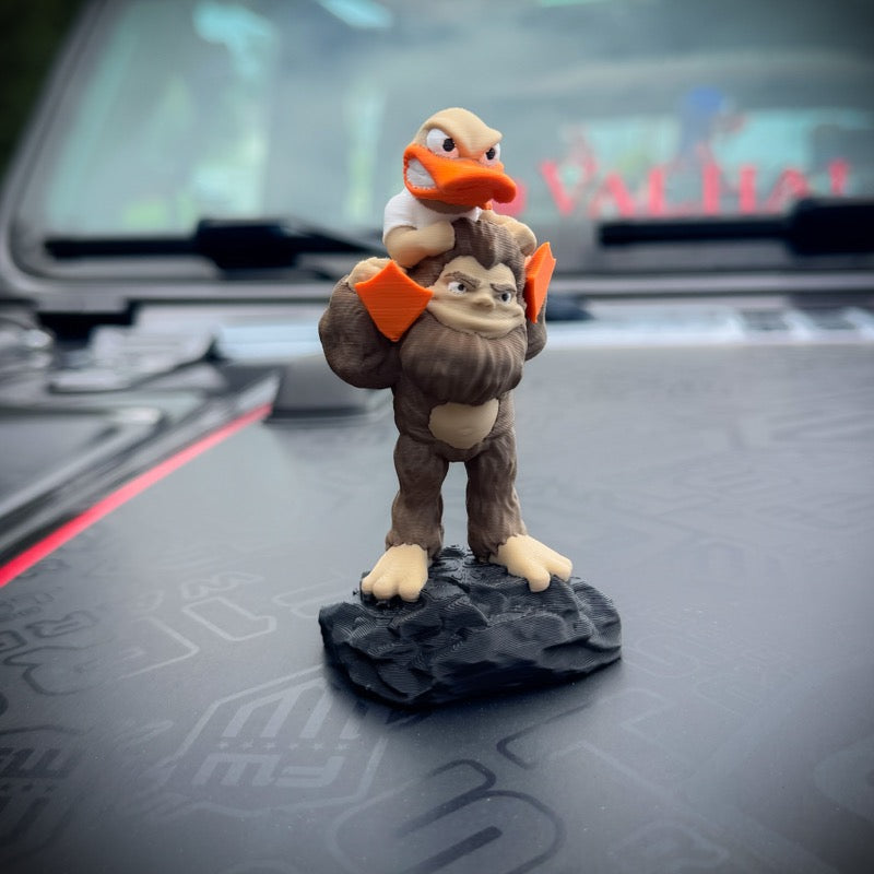 A small figurine of a brown bigfoot carrying a duck on its shoulders, mounted on a dark gray rock base,