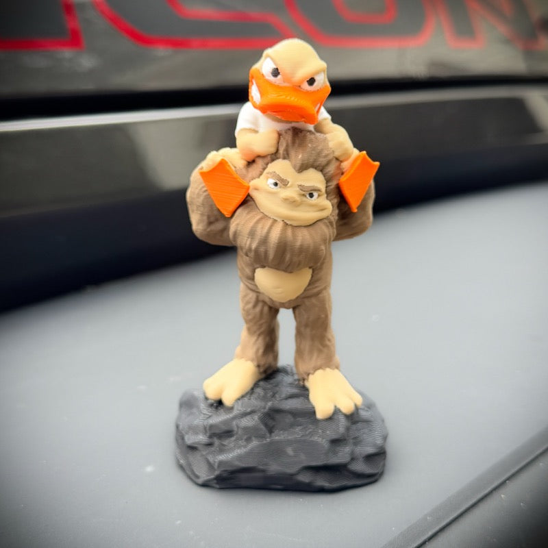 A small figurine of a brown bigfoot carrying a duck on its shoulders, mounted on a dark gray rock base,