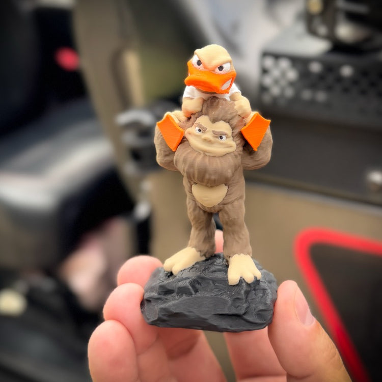 A small figurine of a brown bigfoot carrying a duck on its shoulders, mounted on a dark gray rock base, being held in a person's hand.